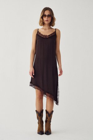 Femeie purtând o rochie tip slip asimetrică de culoare maro închis cu margini de dantelă, ochelari de soare și cizme cowboy bicolore maro, pe un fundal simplu.