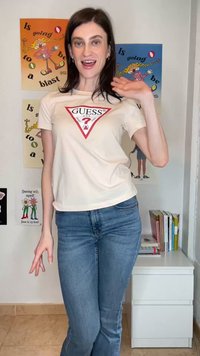 Smilende kvinne med mørkt hår vinker, iført en hvit Guess T-skjorte og blå jeans, stående i et rom med fargerike plakater og et hvitt skap.