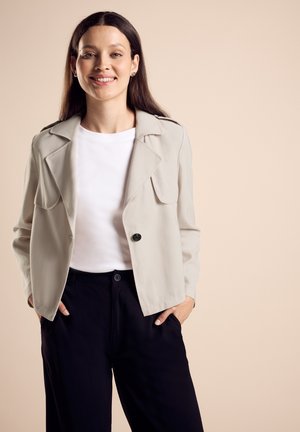 Light beige jacket with a notch collar, front pockets, and single button closure, worn over a white shirt and black pants. Smooth texture.