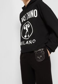 Schwarzer, taillierter Hoodie mit weißem "Moschino Milano"-Logo, kombiniert mit schwarzen Hosen, die ein Strassbären-Design auf der Tasche haben.