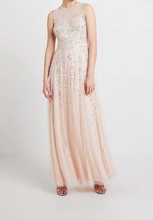 Bez rękawów, długia sukienka w kolorze jasnoróżowym z przezroczystym dekoltem i błyszczącymi zdobieniami z koralików, noszona z sandałami na obcasie z odkrytymi palcami.