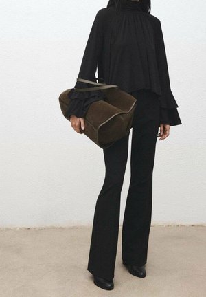 Personne portant un pantalon noir évasé et un haut à manches volantées superposées, tenant un grand sac à main en daim marron contre un mur blanc uni.