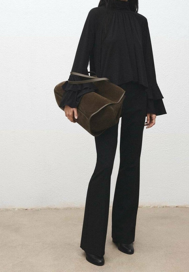 Personne portant un pantalon noir évasé et un haut à manches volantées superposées, tenant un grand sac à main en daim marron contre un mur blanc uni.