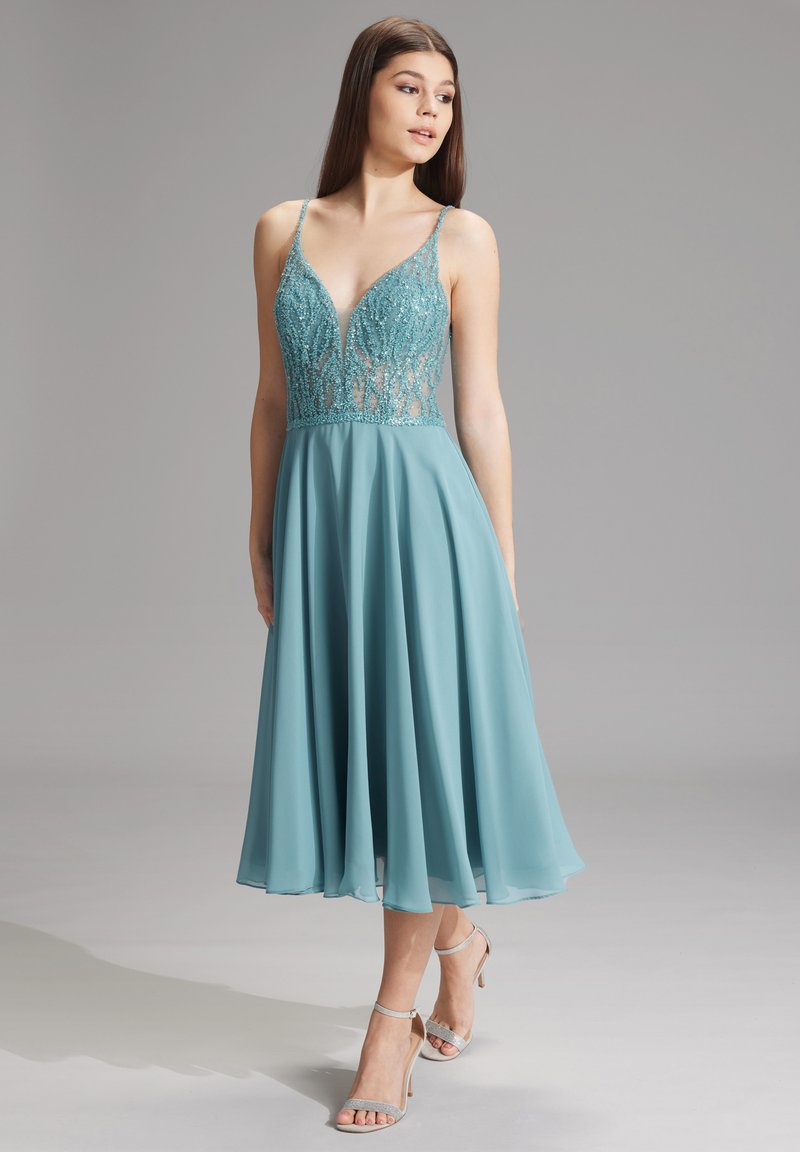 Turkusowa sukienka z głębokim dekoltem w kształcie litery V, haftowaną koronkową górą i zwiewną spódnicą z szifonu, uzupełniona srebrnymi sandałami na obcasie.