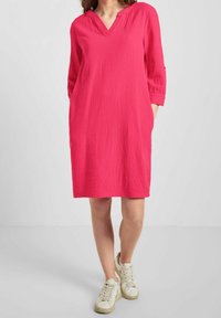 Robe rose à manches trois quarts en tissu texturé avec un décolletté en V et des poches latérales, associée à des baskets blanches.