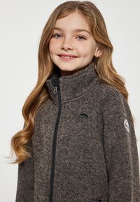 Tamsiai ruda fliso striukė su aukštu užtrauktuku, juodais akcentais ir logotipais ant krūtinės bei rankovės. Minkšta tekstūra ir kasdieniškas dizainas.