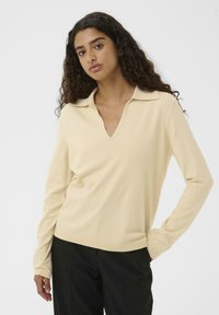 Kvinde med langt krøllet hår iført en lys beige langærmet skjorte med krave og sorte bukser, stående med den ene hånd i lommen mod en hvid baggrund.