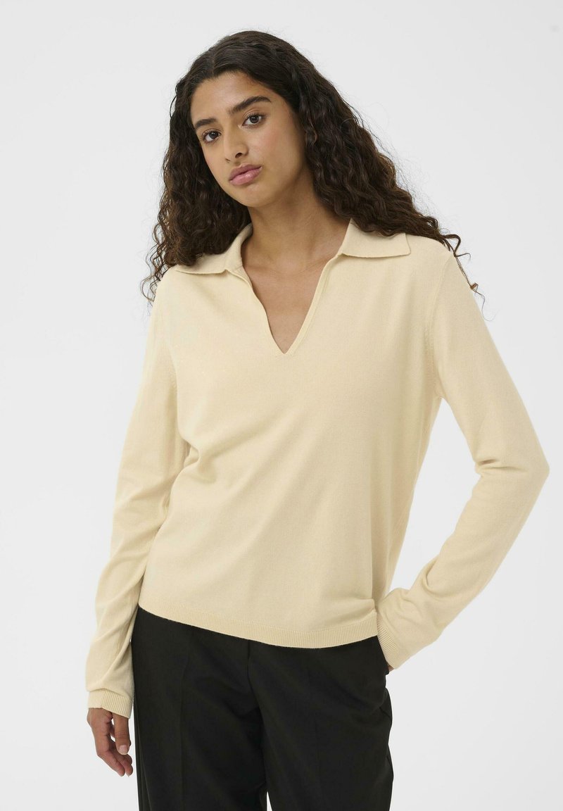 Kvinde med langt krøllet hår iført en lys beige langærmet skjorte med krave og sorte bukser, stående med den ene hånd i lommen mod en hvid baggrund.