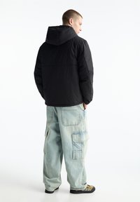 Giacca nera imbottita con cappuccio, caratterizzata da una texture liscia e cuciture. Indossata con jeans larghi azzurri e scarpe slip-on.