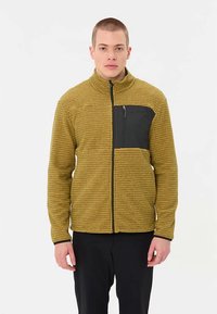 Strukturierte gelb-braune Fleecejacke mit einer schwarzen Brusttasche mit Reißverschluss; hoher Kragen und figurbetonter Schnitt. Schwarze Akzente auf der Brust und den Ärmeln.