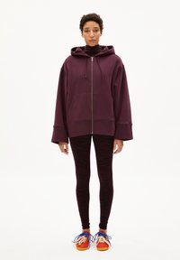 Bordeaux zip-up hoodie met grote voorzak en wijde mouwen, gecombineerd met zwarte leggings met zebra print en multicolore sneakers.