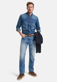 Chemise en denim, bleu clair avec des boutons, associée à un jean bleu décoloré, ceinture en cuir marron et chaussures marron. Veste navy drapée sur le bras.