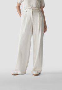 Hochtaillierte, weite Hose aus cremefarbenem Stoff mit Falten und seitlichen Taschen. Kombiniert mit cremefarbenen Schuhen auf einer glatten Fläche.