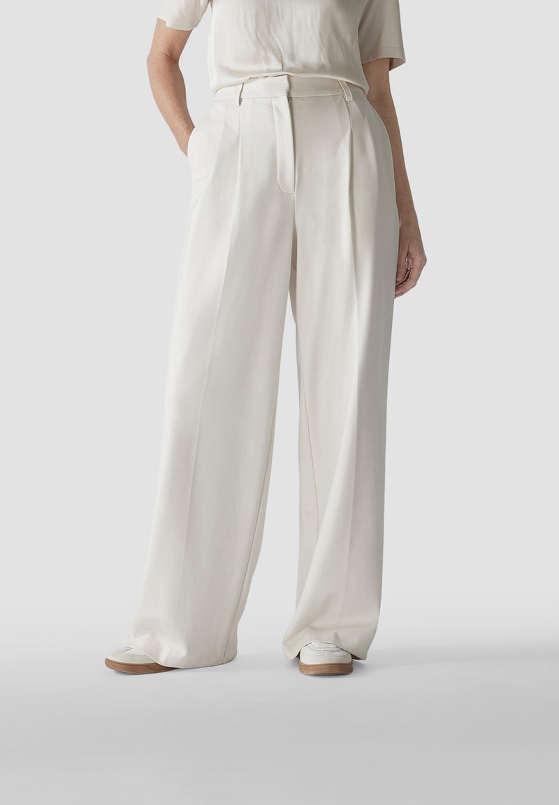 Hochtaillierte, weite Hose aus cremefarbenem Stoff mit Falten und seitlichen Taschen. Kombiniert mit cremefarbenen Schuhen auf einer glatten Fläche.