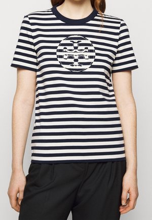 Kvinde iført en kortærmet T-shirt med navyblå og hvide horisontale striber og et centreret cirkulært logodesign, kombineret med sorte bukser.