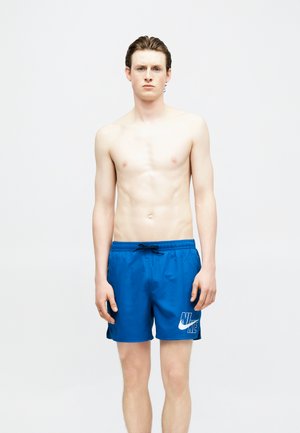 Fiatal férfi rövid barna hajjal, kék Nike úszónadrágot viselve, semleges arckifejezéssel, egyszínű fehér háttér előtt áll.