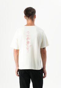 Male model wearing a cream-colored t-shirt with a red floral design on the back, facing away against a plain white background.