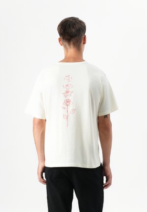 Modello maschile che indossa una t-shirt color crema con un design floreale rosso sulla schiena, girato di spalle su uno sfondo bianco uniforme.