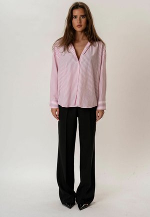 Jeune femme immobile portant une chemise ample à rayures roses, un pantalon large noir et des chaussures noires pointues, sur un fond uni.
