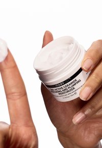 White jar with a black label, containing a thick white cream. Hand applying cream from the jar, with a smooth and glossy texture.