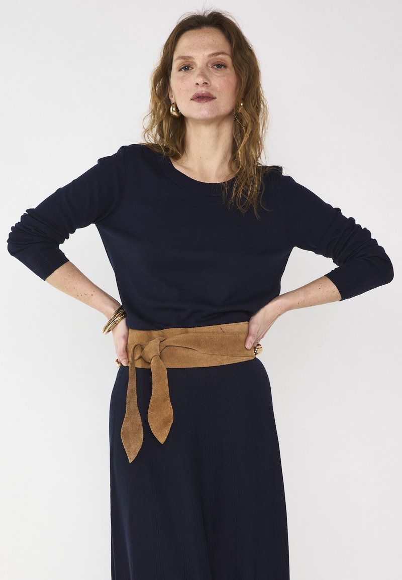 Donna con capelli mossi che indossa una maglia a maniche lunghe blu navy e gonna, cintura larga marrone annodata in vita, orecchini a cerchio dorati e braccialetti, su sfondo bianco.