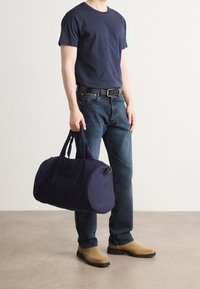 Marineblå duffel bag med to top-håndtag, en rund form og et tekstureret stof. Båret af en person i en marineblå t-shirt, denim jeans og beige støvler.