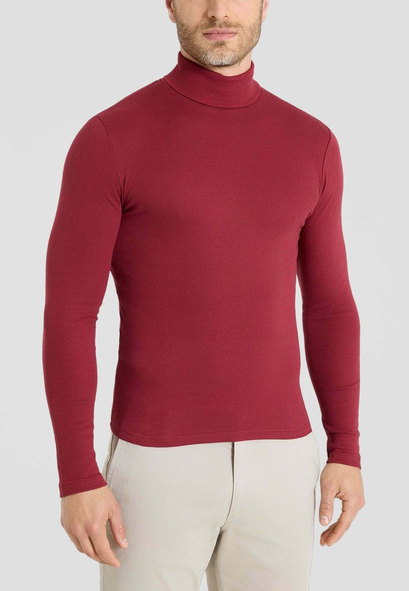 Maglia a maniche lunghe bordeaux con collo alto, dal design aderente, texture morbida, tessuto liscio, abbinata a pantaloni di colore chiaro.