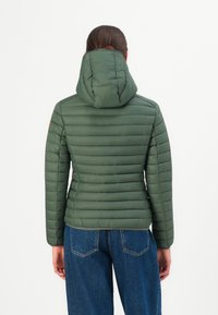 Persoon in een groene gewatteerde jas met capuchon en blauwe jeans, van achteren gezien tegen een effen witte achtergrond.