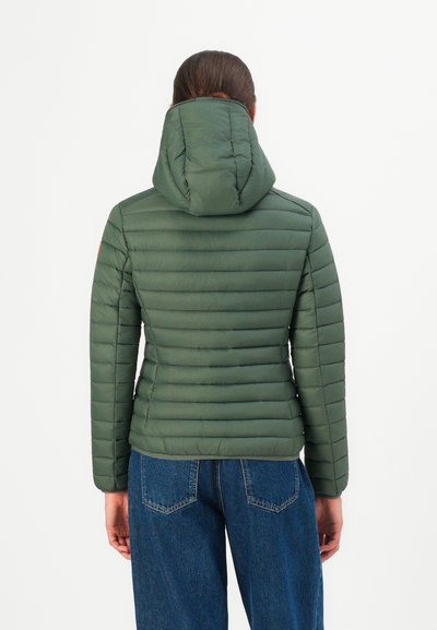 Personne portant une veste matelassée à capuche verte et un jean bleu, vue de dos sur un fond blanc uni.