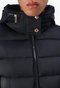 Čierna puzdráková bunda s vysokým golierom, s vreckami na zips a zlatými gombíkmi. Hladká textura s prešívanými detailmi.