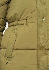 Casaco puffer verde oliva com um design acolchoado, cintura elástica, bolso lateral e fecho de botão de pressão na frente.