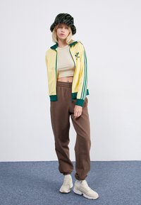 Gul zip-up Adidas träningsjacka med gröna detaljer, beige kort topp, bruna mjukisbyxor och en grön mönstrad bucket hat. Beiga sneakers.