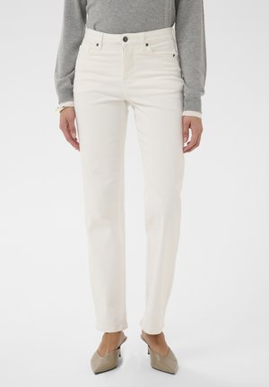 Femme portant un pantalon droit ivoire, un haut à manches longues gris et des escarpins beiges à bout pointu, debout devant un fond blanc.