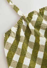 Gros plan d’un tissu texturé à carreaux verts, beiges et blancs avec un bord froncé et une fine lanière.