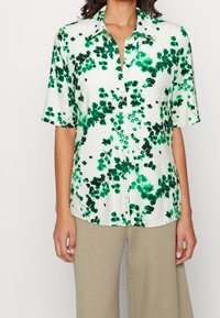 Camicia a maniche corte con bottone, realizzata in tessuto leggero, caratterizzata da un motivo a macchie verdi e nere su uno sfondo bianco.