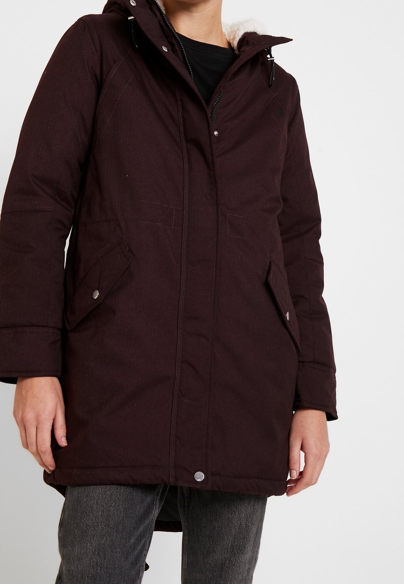 Parka bordeaux scuro con cappuccio, realizzata in tessuto testurizzato, con tasche laterali e chiusura a zip. Il colletto interno è foderato con un morbido tessuto similpelliccia.