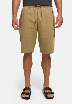 Uomo che indossa pantaloncini beige con coulisse e tasche con cerniera e sandali neri con fibbia, in piedi davanti a uno sfondo bianco semplice.