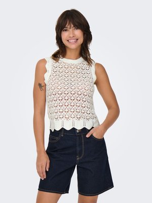 Femme souriante portant un haut blanc en maille sans manches avec des bords festonnés et un short en jean foncé longueur genou, la main dans la poche.