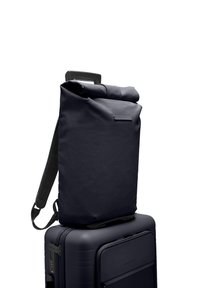 Sac à dos roll-top bleu marine en tissu texturé, avec des bretelles ajustables et un profil mince, reposant sur une valise rigide assortie.