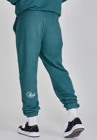 Teal sweatpants med avslappnad passform, elastiska manschetter, sidofickor och vitt logomotiv på underbenet. Materialet verkar mjukt och texturerat.