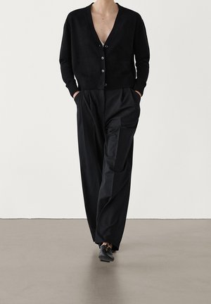 Osoba ubrana w czarny, zapinany na guziki kardigan, luźne czarne spodnie z kieszeniami oraz czarne sznurowane buty, stojąca na jednolitym tle.