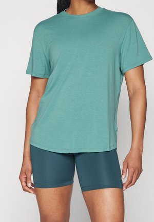 Femme portant un t-shirt ample bleu sarcelle à manches courtes et un cuissard ajusté bleu sarcelle foncé, debout devant un fond uni.