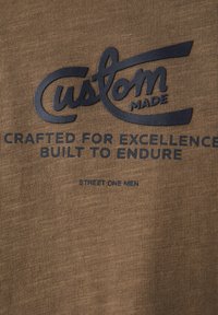 Hnědý látkový tričko s tmavým potiskem textu "vyrobeno na míru," "vytvořeno pro dokonalost," a "postaveno, aby vydrželo" s texturovanými detaily.