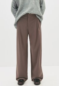 Personne portant un pantalon marron large, un pull gris et des chaussures en cuir noir se tenant contre un fond uni.