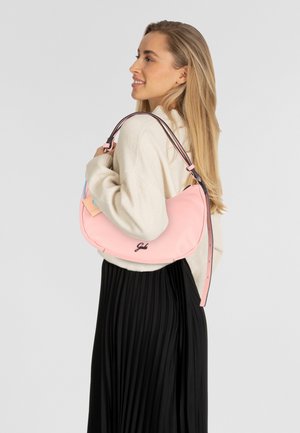 Roze leren schoudertas met een gebogen ontwerp, een bicolor schouderriem en logo-details. Het model draagt een crèmekleurige trui en een zwarte geplooide rok.