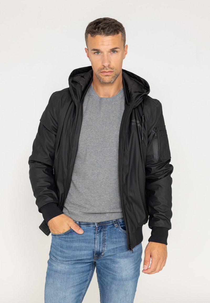 Czarna bomberka z kapturem, z zamykanymi kieszeniami, elastycznymi mankietami i gładką fakturą. Nosi się ją na szarym T-shircie i niebieskich dżinsach.