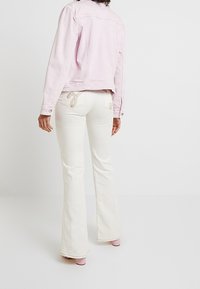 Kobieta w jasnoróżowej dżinsowej kurtce i białych dzwonowatych spodniach z haftem na kieszeniach, stojąca na tle jasnego, jednolitego tła.