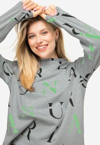 Szara bluza wykonana z miękkiego materiału, z mieszanką czarnych i zielonych liter w różnych rozmiarach rozłożonych po powierzchni. Rękawy są wydłużone.