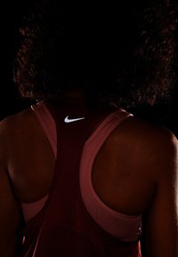 Bordowy sportowy top na ramiączkach z wycięciem na plecach w kształcie racerback, z subtelnym białym logo Nike na górnej części pleców. Powierzchnia wydaje się gładka.