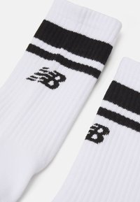 Calcetines blancos de canalé con una raya negra en la parte superior. Presentan un logo negro de New Balance en el lateral. Textura suave y altura media.